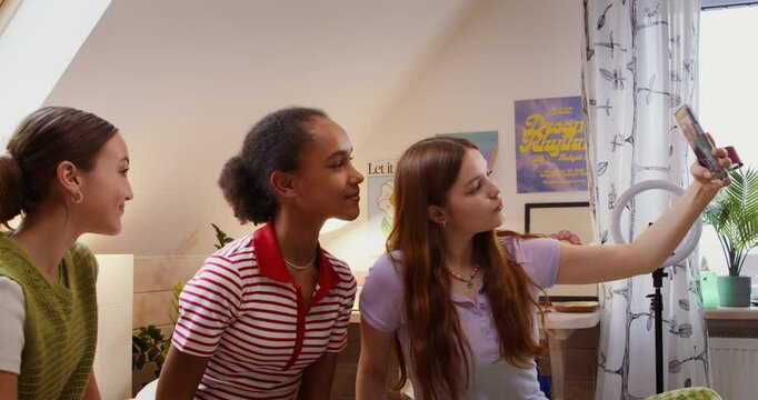 Three cute girls are taking selfies on a mobile phone. They're sitting on the bed at home.