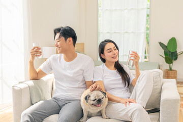 Happy Asian family couple drinking a glass of pure water and playing with senior Pug dog in living...