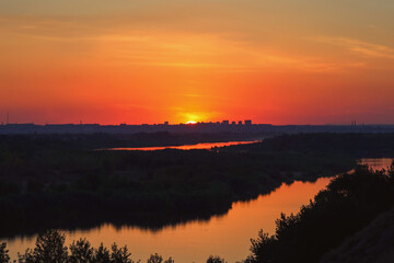 Fototapeta premium The setting sun paints the sky with fiery hues of orange and red, casting a warm glow over the silhouette of a cityscape and a tranquil river meandering through lush vegetation