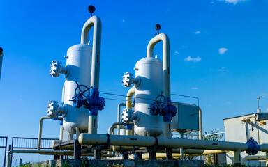Two large gas processing units with valves and piping stand prominently against a bright blue sky at a manufacturing site during the day
