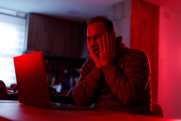 Man reacts with frustration while working on laptop in dimly lit room