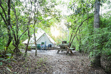 A canvas tent surrounded by lush forest and morning light creates a peaceful campsite scene. Includes chairs and a picnic table for a full camp setup.