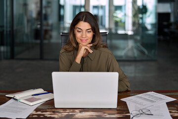 Smiling busy mature middle aged professional business woman, female company manager executive or entrepreneur looking at laptop computer in office working on online financial data sitting at desk.