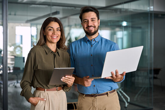 Team of two happy business partners man and woman standing in office looking at camera. Portrait of 2 company managers, corporate executive professionals, businessman and businesswoman at work.