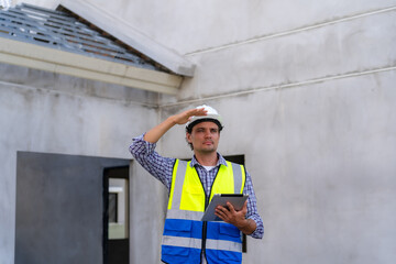 Young construction engineer looking forward with determination wearing hard hat and using digital tablet while working on construction site