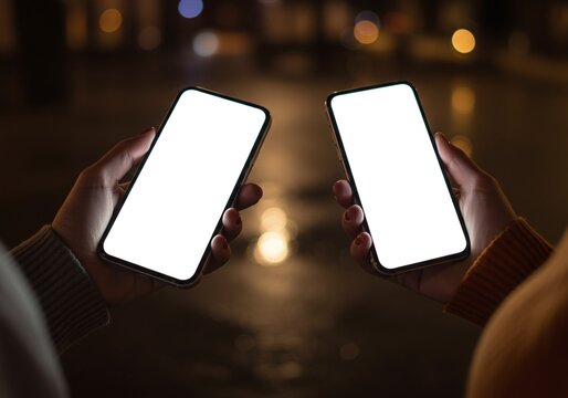 Photo of Two people holding smartphones with blank screens at night in a city, showcasing modern technology and communication against a blurred background of city lights - Powered by Adobe