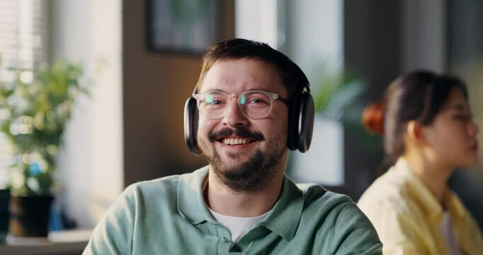 A man wearing headphones looks at the camera. He talks and laughs while sitting next to his colleague.