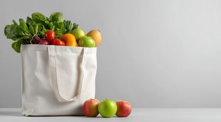 A white tote bag brimming with fresh produce; spinach, beets, tomatoes, peppers, apples, and mango, sits on a table against a grey backdrop. Three additional apples rest beside the bag