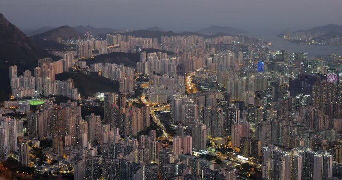 Night view of Hong Kong dense cityscape from above, featuring tall residential towers of Kowloon and warmly lit streets below. Soft evening afterglow lingers in sky, not yet fully dark