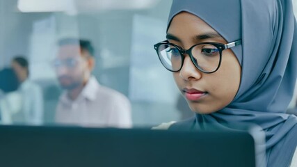 Young Asian woman in hijab with eyeglasses in front of laptop monitor in office. A serene young woman in hijab collaborates effectively on her laptop. - Powered by Adobe