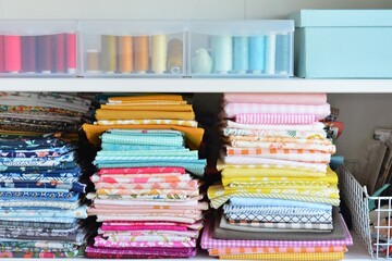 Stacks of bright cotton fabrics, sewing threads in plastic boxes, blue paper box and white metal small basket with scrapbooking stuff on shelves