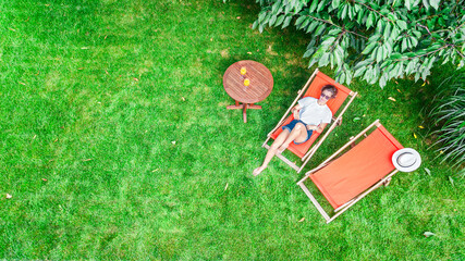 Young girl relaxes in summer garden in sunbed deckchair on grass, woman reading book outdoors in green park on weekend, aerial drone view from above