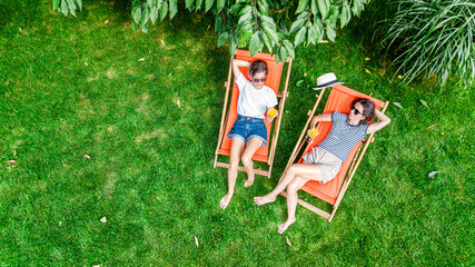 Young girls relax in summer garden in sunbed deckchairs on grass, women friends have fun outdoors in green park on weekend, aerial drone view from above