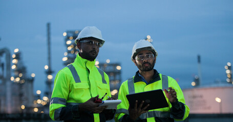  Two male engineers or technicians wearing white hard hats and high-visibility vests, working with digital tablet outdoors front of factory at night.