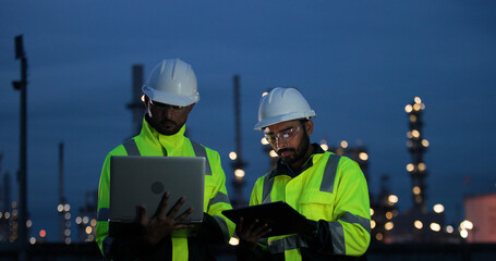 Fototapeta premium Two male engineers or technicians wearing white hard hats and high-visibility vests, working with digital tablet outdoors front of factory at night.