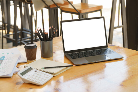Mockup image of laptop computer screen with blank white background for advertising text on a large wooden table with office supplies at a bright modern workplace. Marketing and business concept.