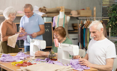 Elderly men and women sew on sewing machine and discussing pattern in group during lesson