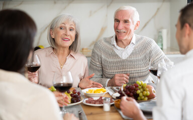 Merry old parents having lunch with their son and his girlfriend while drinking wine and talking warmly in their house