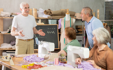 In sewing training workshop, friendly elderly male instructor standing at blackboard with chalk,...