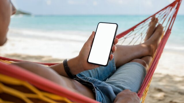 Man relaxing in hammock on beach holding smartphone with blank screen, concept for travel app, mobile marketing, and summer vacation promotion