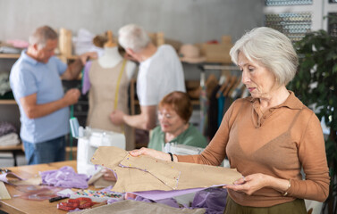 Focused elderly woman reviewing paper pattern pinned to fabric surrounded by enthusiastic classmates working on different sewing tasks in training tailoring workshop for older adults..