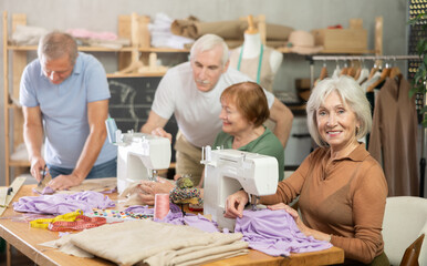 Fototapeta premium Older woman smiling while working at sewing machine during hands-on course for seniors, surrounded by classmates preparing patterns, fabric and fitting garments