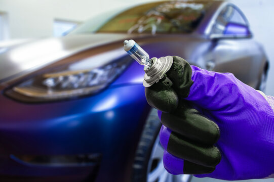 Mechanic wearing protective gloves holds a halogen car headlight bulb near the front of a modern car. Concept of car maintenance or bulb replacement.