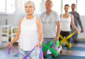 Elderly woman kneeling performs exercises with elastic band during Pilates workout. Bring together...