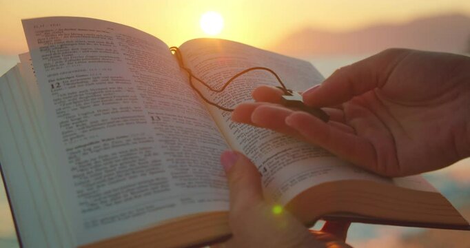 Hands hold an open Bible and a cross pendant at the beach during a sunset as the light shines over the pages