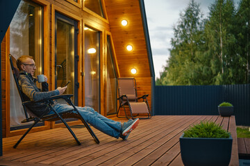 Man sits with a cup of tea in the evening on the terrace illuminated by outdoor wall lamps