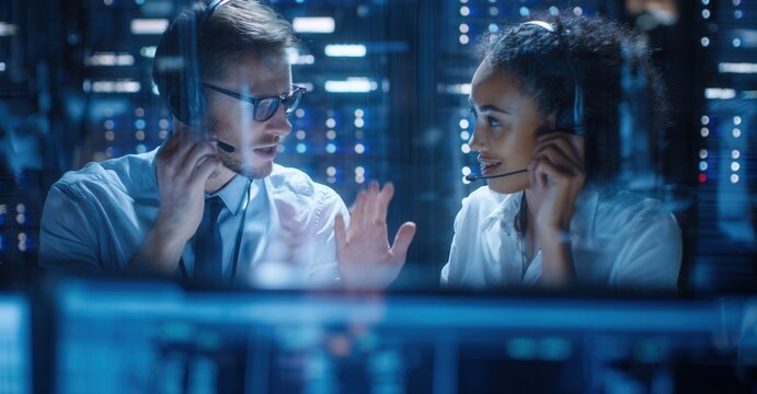 Two technicians wearing headsets communicate in a server room, overseeing system operations and providing technical support in a high-tech environment.