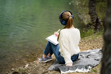 Rear view of woman writing in notebook outdoors journaling keeping diary studying listening music resting relaxing alone wearing headphones, white sweater and jeans sitting by lake in spring park
