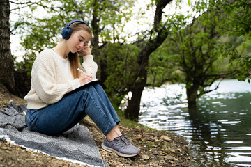 Full body of calm thoughtful woman writing in notebook keeping diary relaxing study listening music...