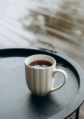 A white mug of hot coffee on a black metal table outdoors. Cozy and peaceful moment on a rainy day.