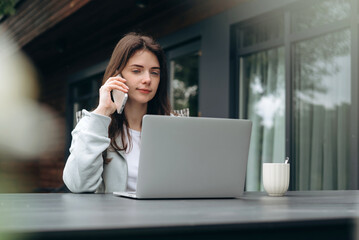 Young woman working on laptop and talking on phone outdoors. Freelancer, remote work, and casual lifestyle concept.