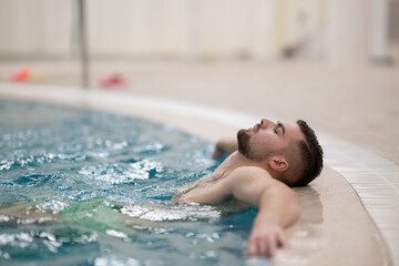 Man floating peacefully in warm spa water with eyes closed and arms outstretched on the edge of the pool.