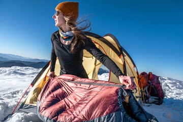 Young woman removing a sleeping bag from a tent in snowy mountain landscape, winter camping gear in focus © XArt
