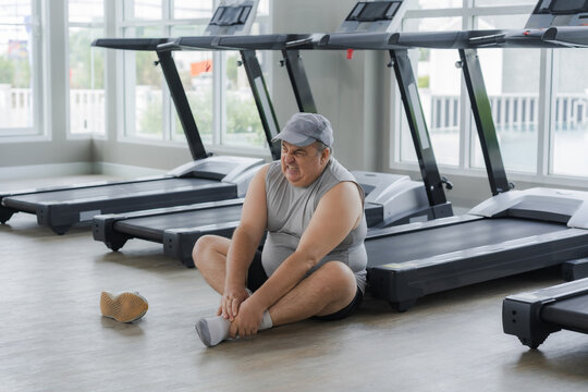 Fat man sits with his hand on his ankle after injuring his ankle while running on a treadmill in a gym.
