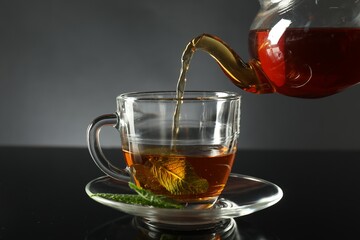 Pouring freshly brewed tea from teapot into cup at dark mirror table, closeup