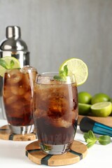Tasty Cuba Libre cocktail in glasses, ingredients, muddler and shaker on white wooden table