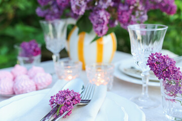 Beautiful lilac flowers, zephyrs, plates, cutlery, glasses and burning candles on table in garden, closeup