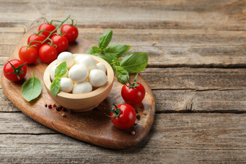 Tasty mozzarella cheese balls in bowl, basil, peppercorns and tomatoes on wooden table, space for text