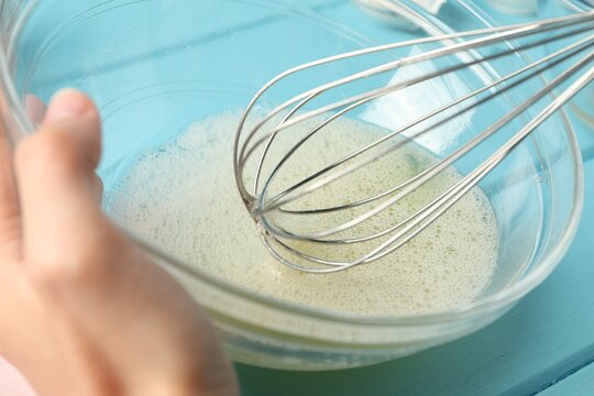 Woman whisking egg whites in bowl at light blue table, closeup