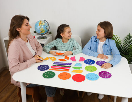 Smiling teacher teaches two children with colorful fraction circles and time clocks during a math lesson. The educational activity promotes hands-on learning and student engagement at home.
