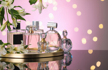 Many different perfume bottles and flowers on mirror surface against pink background with blurred...