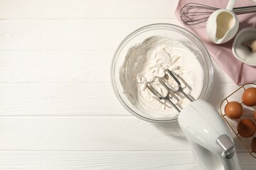 Hand mixer with whipped cream, eggs and other products on white wooden table, flat lay. Space for text