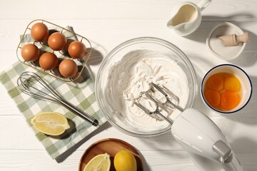 Hand mixer with whipped cream, eggs and other products on white wooden table, flat lay