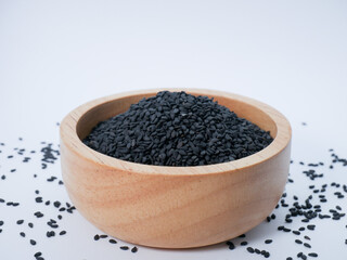 Wooden bowl of black sesame seeds with some scattered around, presented on a bright surface.