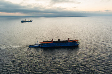 Drone aerial in the Baltic Sea: a tug pulls a barge in the foreground, a cargo ship on the horizon at dawn.