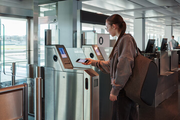 A young woman uses her smartphone to scan a boarding pass at an automated airport gate. Concept of digitalization, self-check-in and independent travel.